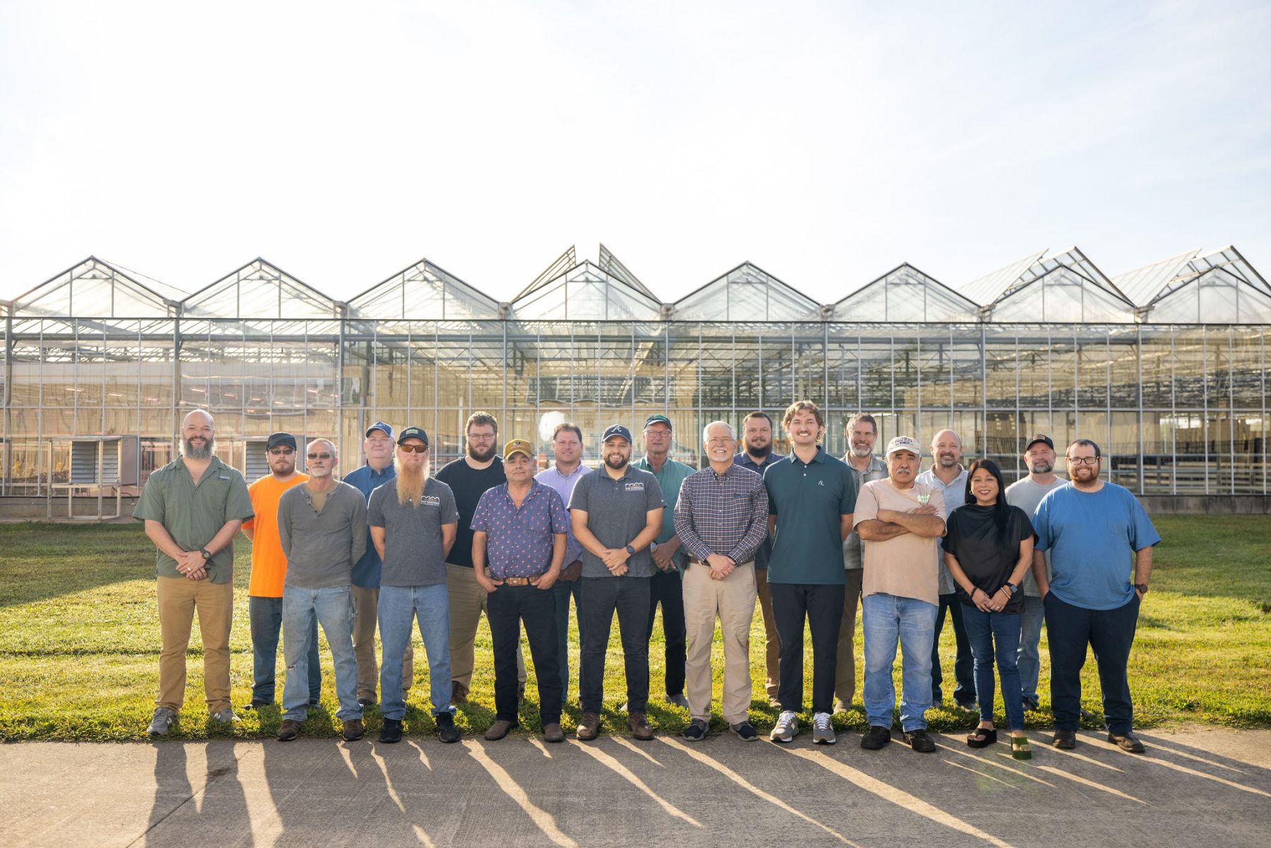 A group of sixteen people stand in a row outside in front of a large greenhouse structure on a sunny day. The group is standing on grass and pavement, facing the camera. - Van Wingerden Commercial Greenhouses