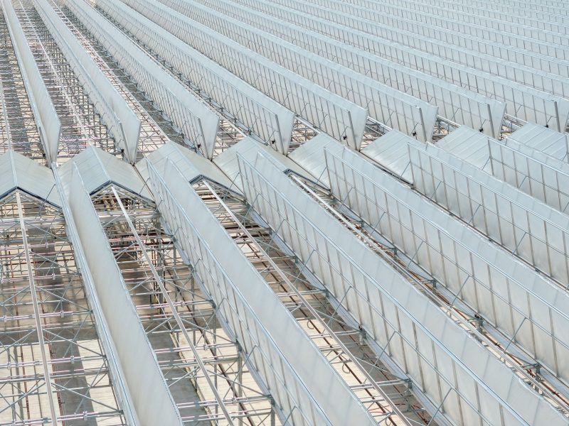 Rows of large, metallic, angled structures are arranged in a repeating pattern. Scaffolding and support beams are visible beneath the structures, creating a geometric, industrial appearance. - Van Wingerden Commercial Greenhouses
