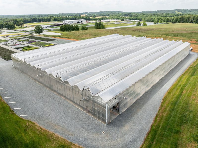 A large, modern greenhouse with a transparent roof sits on a gravel lot, surrounded by grassy fields and a few smaller buildings in the distance under a cloudy sky. - Van Wingerden Commercial Greenhouses
