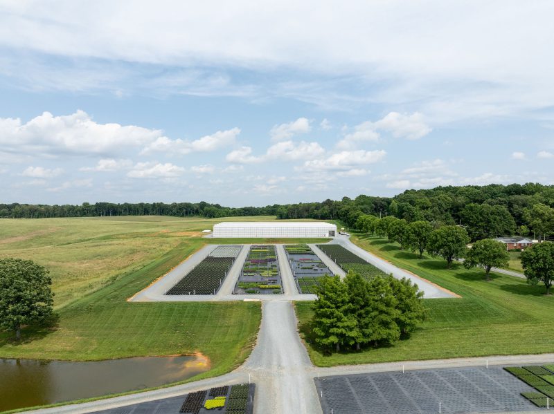 Aerial view of a cultivated agricultural area featuring organized rows of crops, a greenhouse in the background, gravel roads, grassy fields, and clusters of trees under a partly cloudy sky. - Van Wingerden Commercial Greenhouses