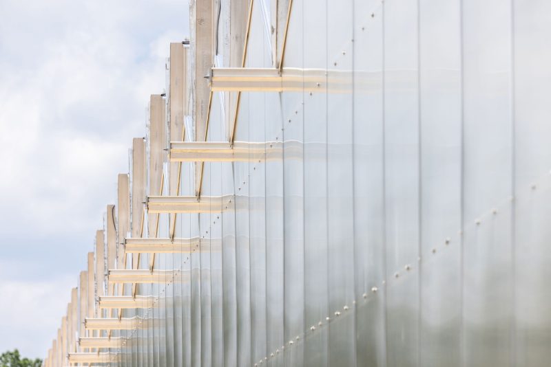Close-up view of a modern building facade with vertical metal beams and translucent glass panels, photographed from an angle that highlights repetition and reflection under a partly cloudy sky. - Van Wingerden Commercial Greenhouses