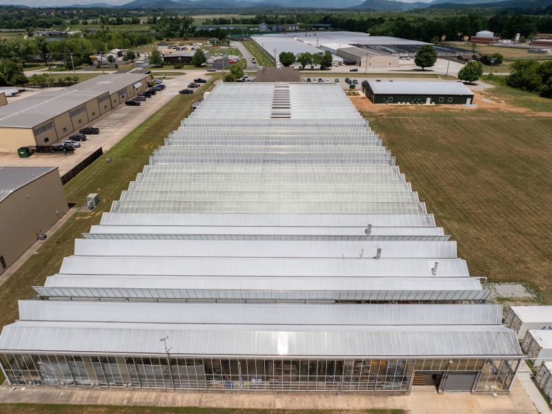 Aerial view of a large greenhouse complex with multiple parallel-roofed sections, surrounded by industrial buildings, parking lots, grassy areas, and distant hills. - Van Wingerden Commercial Greenhouses