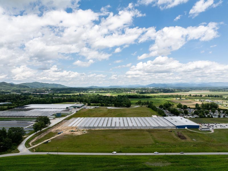 Aerial view of a large industrial building with a white roof, surrounded by green fields, trees, and roads. Several cars are parked near the building, and hills and clouds are visible in the background. - Van Wingerden Commercial Greenhouses