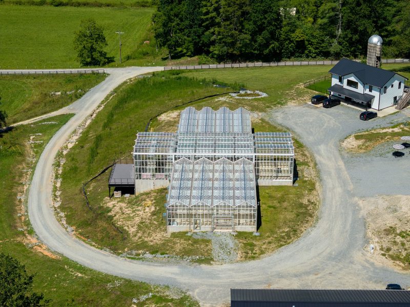 Aerial view of a greenhouse complex with multiple connected glass structures, surrounded by a gravel road and grassy areas. A white farmhouse with a silo stands nearby. Trees and open fields border the property. - Van Wingerden Commercial Greenhouses