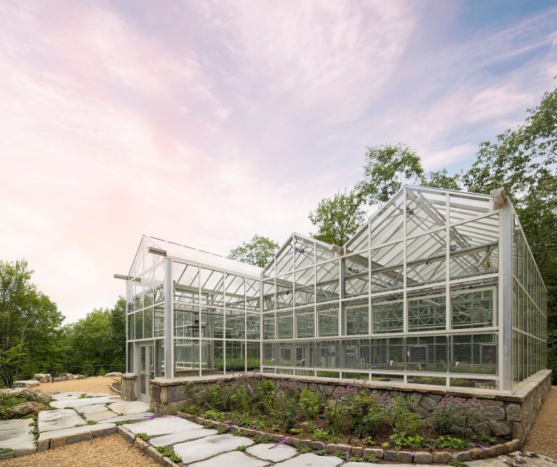 A large, modern glass greenhouse with a metal frame sits on a stone foundation, surrounded by trees and a landscaped stone walkway under a partly cloudy sky. - Van Wingerden Commercial Greenhouses