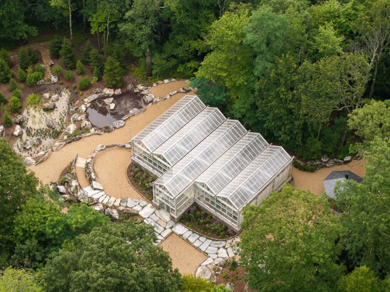 A large greenhouse with a glass roof is surrounded by trees, stone paths, and landscaped garden beds in an aerial view. The surrounding area includes rocks, gravel walkways, and patches of greenery. - Van Wingerden Commercial Greenhouses