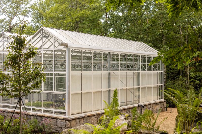 A white, glass-paneled greenhouse stands on a stone foundation surrounded by trees, greenery, and a gravel path. The greenhouse has a peaked roof and several plants visible inside. - Van Wingerden Commercial Greenhouses