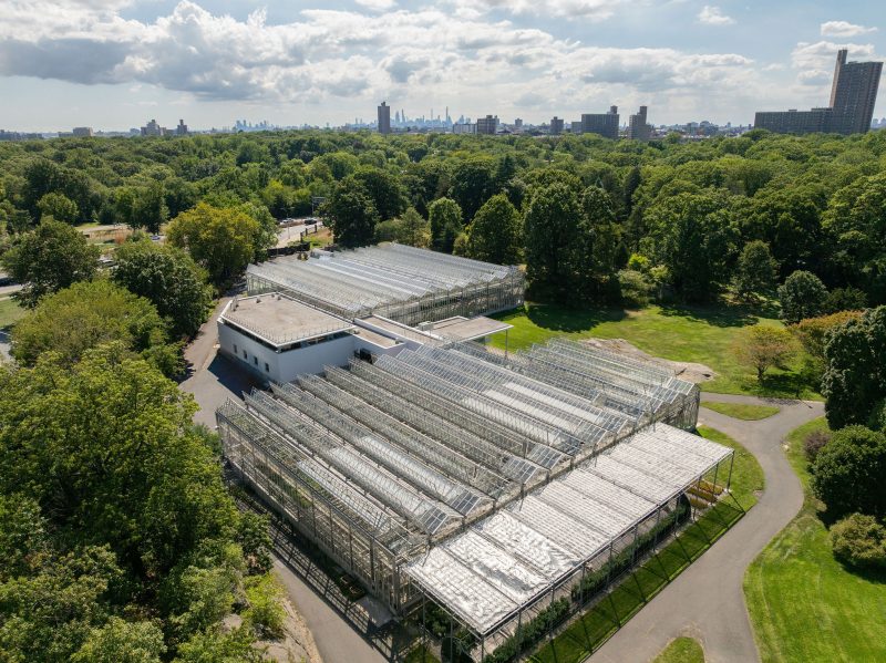 Aerial view of two large glass greenhouses next to a white building, surrounded by trees and greenery, with a city skyline visible in the background under a partly cloudy sky. - Van Wingerden Commercial Greenhouses