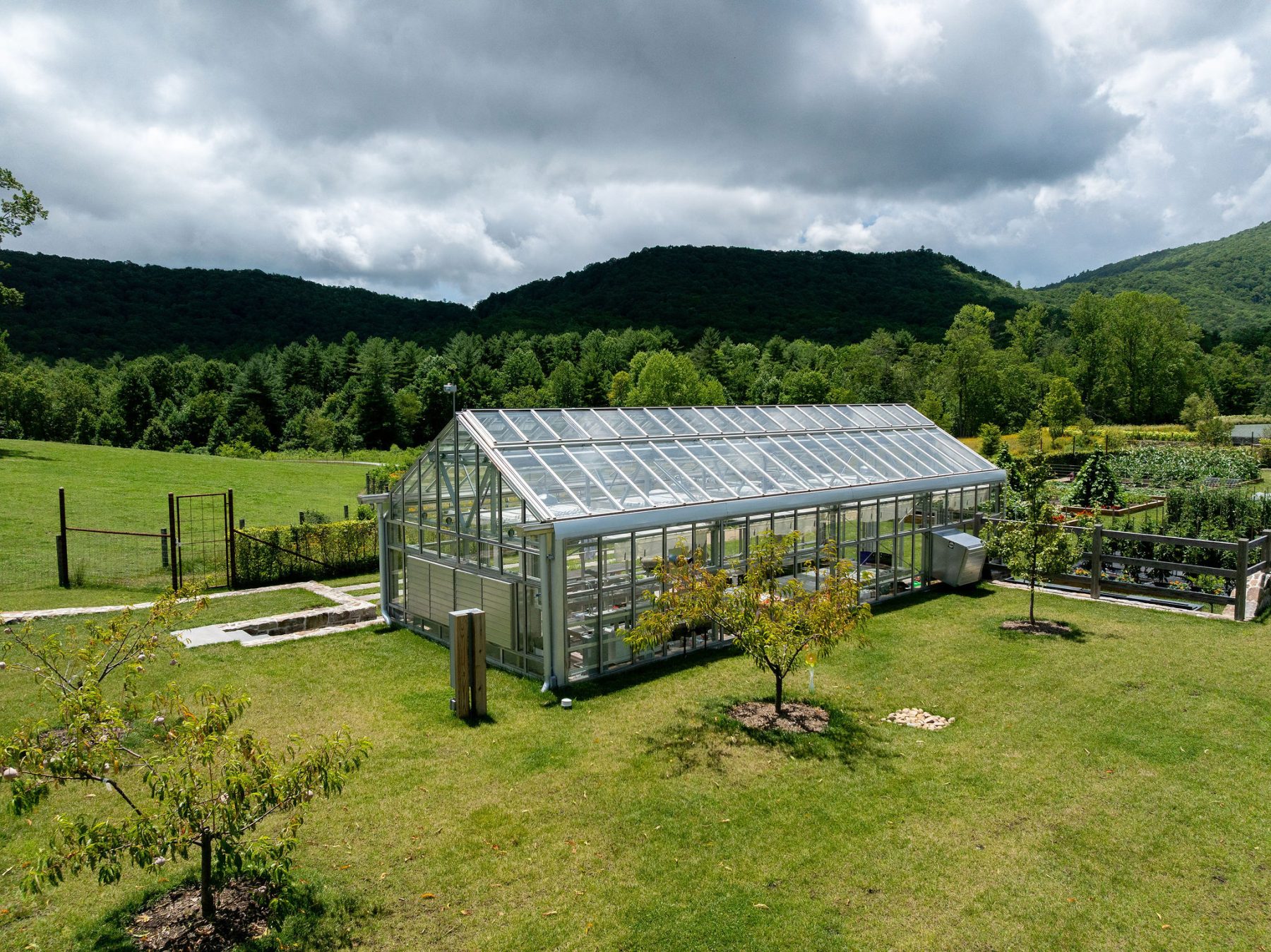 A glass greenhouse stands on a grassy lawn surrounded by young trees, with forested hills and a cloudy sky in the background. Raised garden beds and a fence are visible nearby. - Van Wingerden Commercial Greenhouses