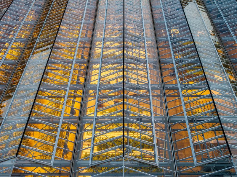 A close-up view of a modern buildings glass facade, featuring repeating geometric patterns and metal beams, with warm yellow reflections visible on the glass panels. - Van Wingerden Commercial Greenhouses