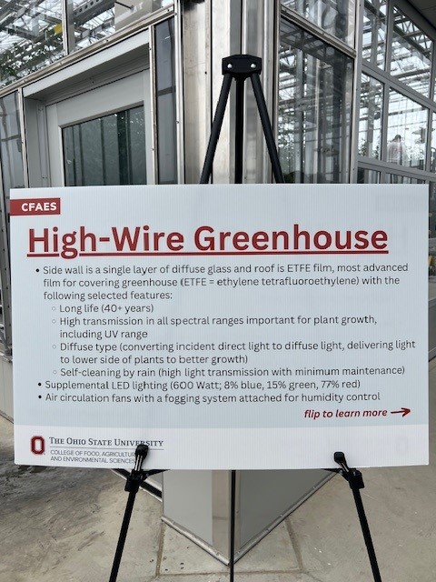 A sign on an easel in a greenhouse describes features of a High-Wire Greenhouse, including ETFE film use, lighting, self-cleaning, and climate controls. The sign is from The Ohio State University CFAES. - Van Wingerden Commercial Greenhouses