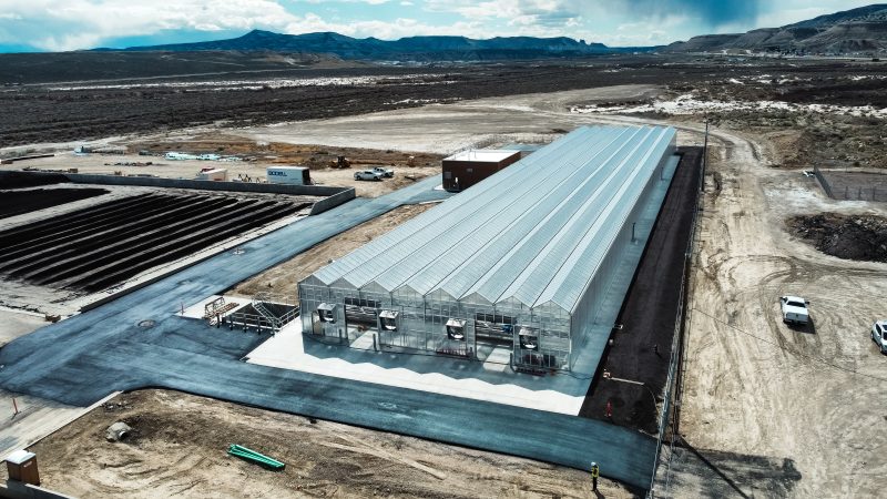 Aerial view of a large, modern greenhouse building on a paved lot surrounded by dirt and open land, with mountains visible in the background. Construction materials and vehicles are scattered around the site. - Van Wingerden Commercial Greenhouses